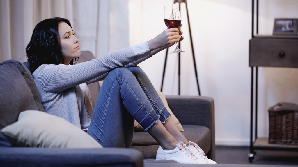 mujer deprimida en jersey beige y jeans sentados en el sofá y sosteniendo una copa de vino tinto en manos extendidas en casa - Foto, imagen