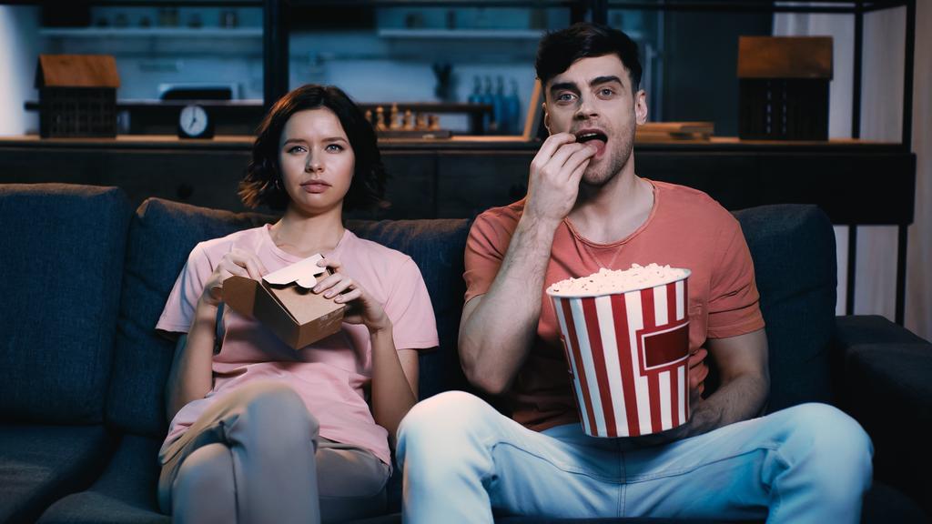 man eating popcorn near girlfriend holding cardboard box with chinese food while watching movie - Photo, Image