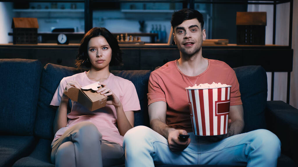onnellinen mies tilalla ämpäri popcornia ja klikkaamalla kanava lähellä tyttöystävä tilalla pahvilaatikko kiinalaista ruokaa  - Valokuva, kuva