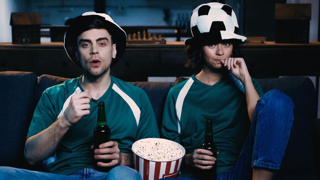підкреслені фанати футболу в капелюхах, що тримають пляшки пива і спостерігають за чемпіонатом у вітальні
  - Фото, зображення