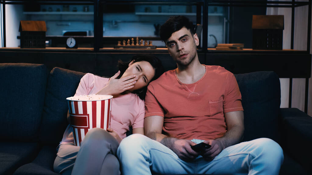tired woman holding popcorn bucket and yawning while leaning on shoulder of boyfriend and watching movie  - Photo, Image