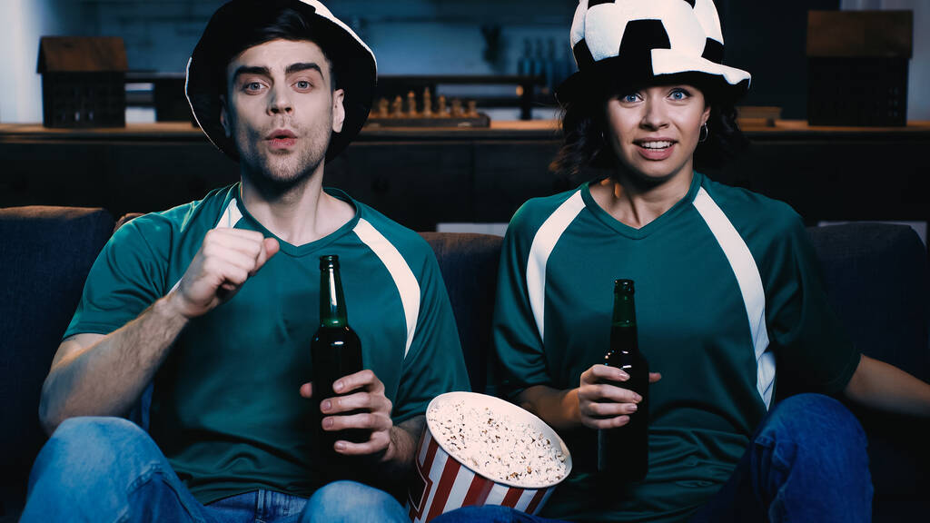 pareja estresada en abanicos sosteniendo botellas de cerveza y viendo el campeonato cerca de cubo con palomitas de maíz  - Foto, Imagen
