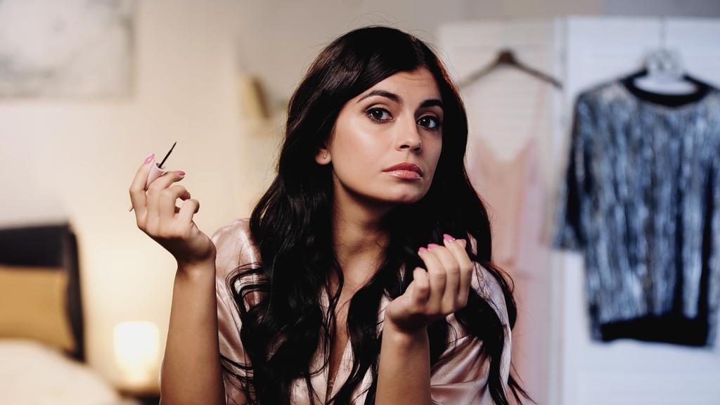 young woman in pink silk peignoir doing makeup with eyeliner in bedroom - Photo, Image