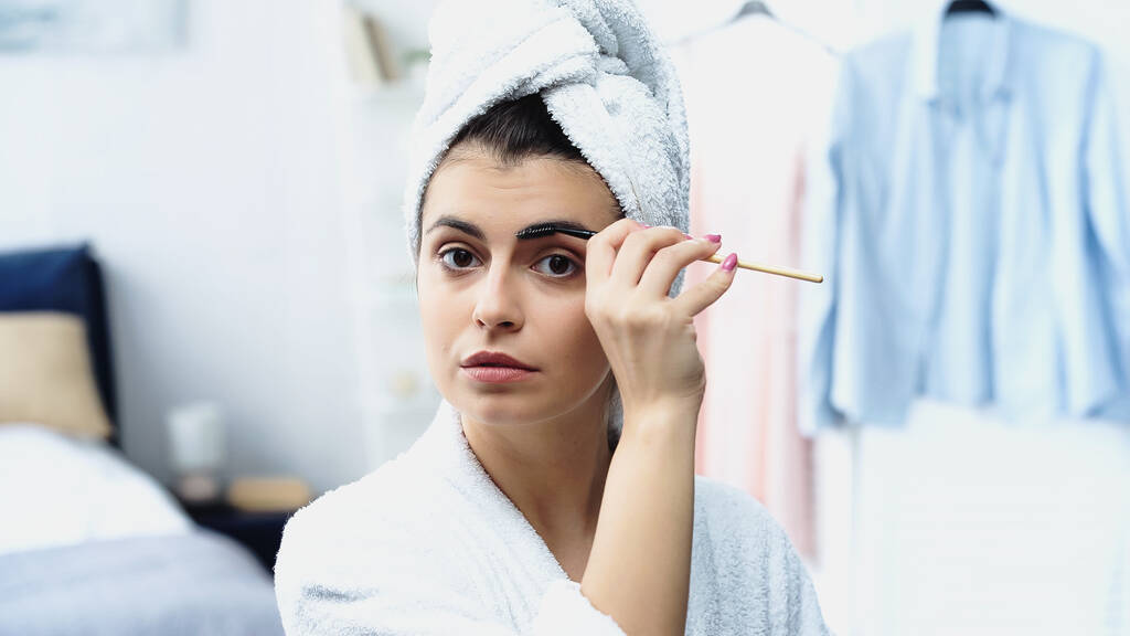 young woman with head wrapped in towel styling eyebrow with cosmetic brush in bedroom - Photo, Image