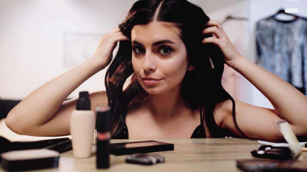brunette nuori nainen säätämällä hiukset lähellä pöytää koriste kosmetiikka makuuhuoneessa - Valokuva, kuva
