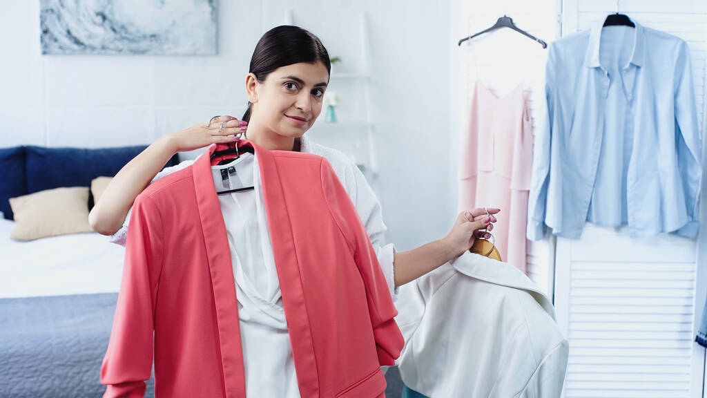 smiling brunette young woman in bathrobe holding clothes on hangers in bedroom  - Photo, Image