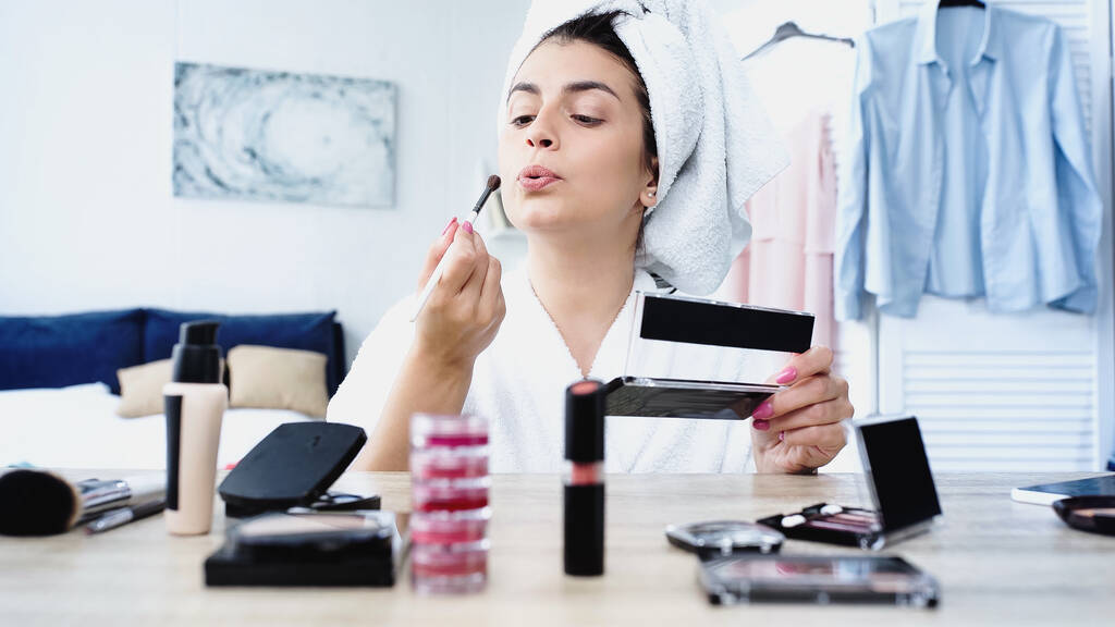 young woman in bathrobe blowing on cosmetic brush near table with decorative cosmetics in bedroom - Photo, Image