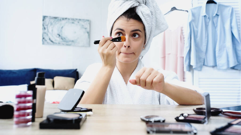 young woman with head wrapped in towel grimacing and applying face foundation with cosmetic brush in bedroom - Photo, Image