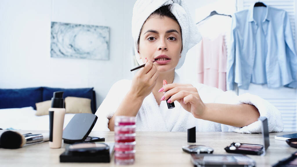 mujer joven con la cabeza envuelta en toalla aplicando lápiz labial con cepillo cosmético en el dormitorio - Foto, Imagen