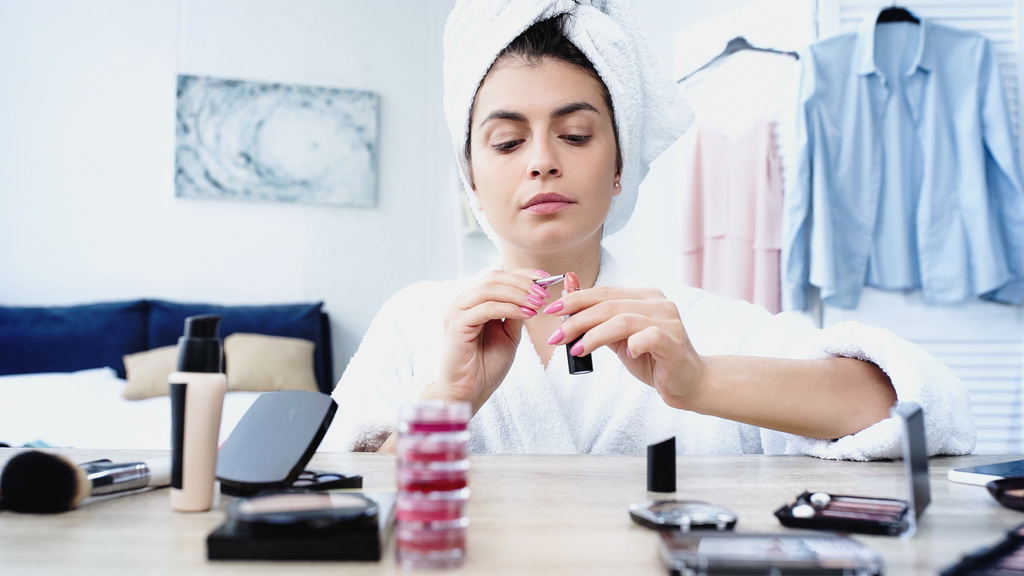 mujer joven en albornoz con cabeza envuelta en toalla aplicando lápiz labial con cepillo cosmético en el dormitorio - Foto, Imagen