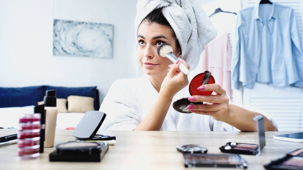 giovane donna in accappatoio con testa avvolta in asciugamano applicando cipria con spazzola cosmetica in camera da letto - Foto, immagini