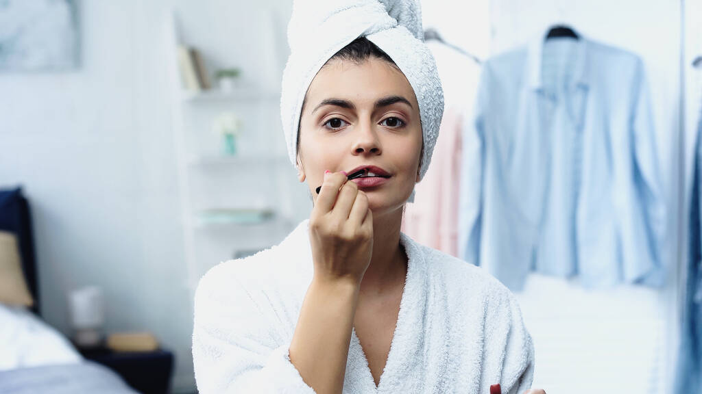 mujer joven en albornoz con cabeza envuelta en toalla aplicando lápiz labial con cepillo cosmético en el dormitorio - Foto, imagen