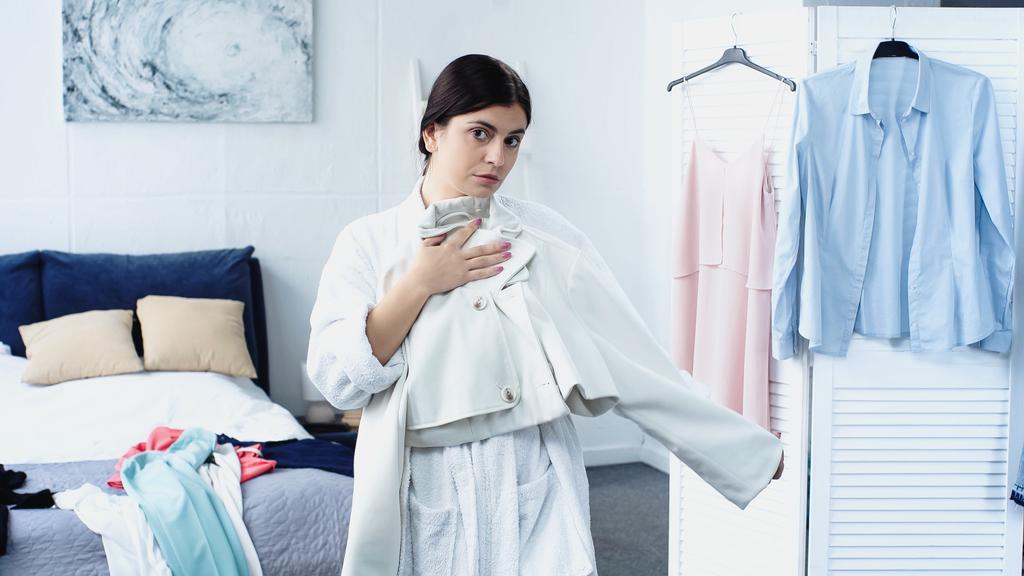 Nuori nainen valkoisessa kylpytakissa sopiva takki sängyn lähellä vaatteita makuuhuoneessa - Valokuva, kuva