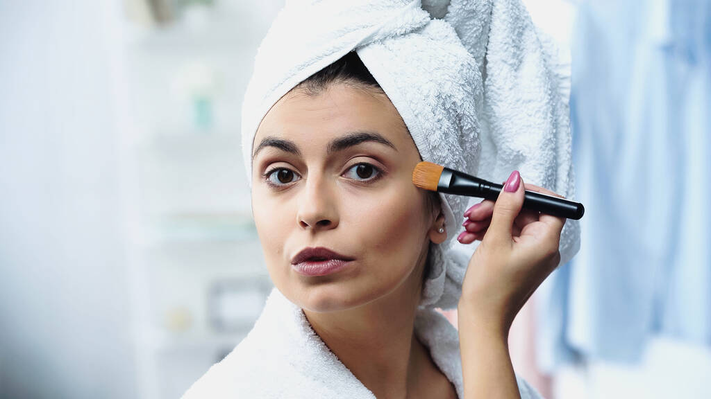 mladá žena s hlavou zabalenou v ručníku nanášení nadace na obličej s kosmetickým štětcem v ložnici - Fotografie, Obrázek