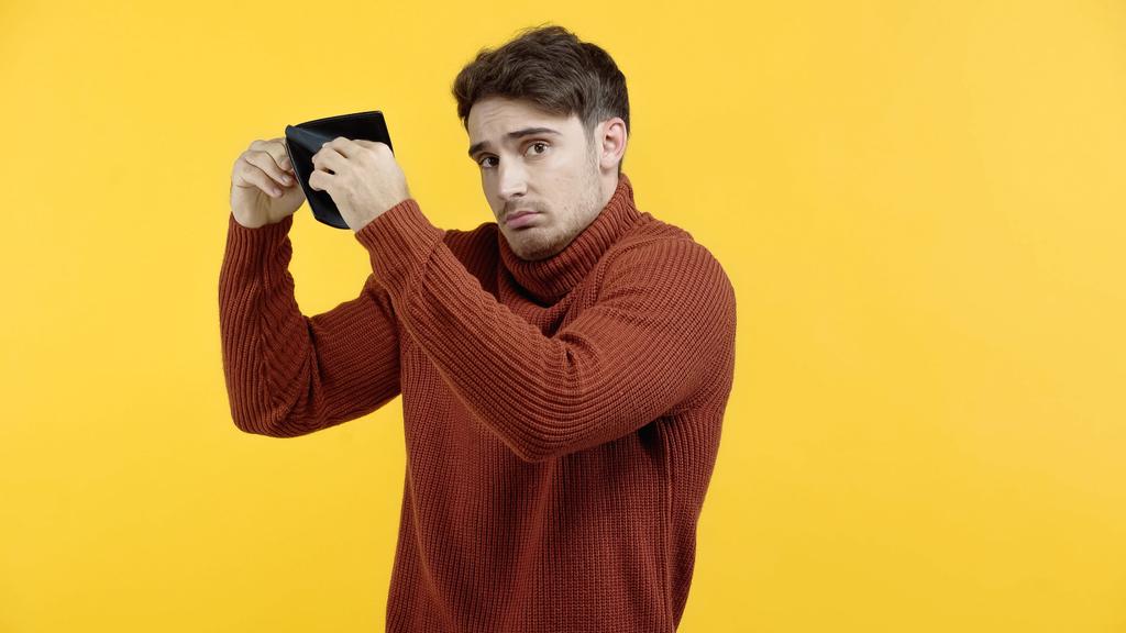 Homem descontente em suéter segurando carteira vazia isolada no amarelo  - Foto, Imagem