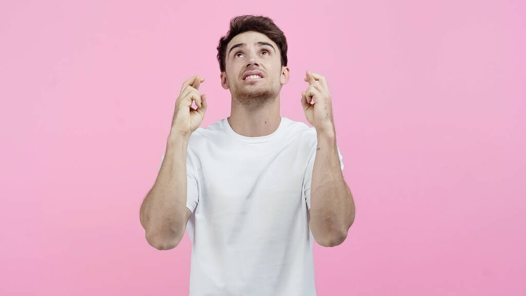 Hombre joven en camiseta blanca mostrando los dedos cruzados y mirando hacia arriba aislado en rosa - Foto, Imagen