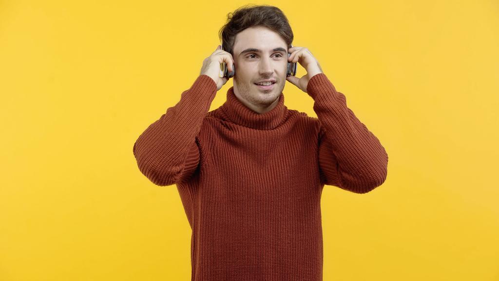 Süveter giyen genç adam kulaklıkla müzik dinliyor ve sarıda izole bir şekilde gülümsüyor.  - Fotoğraf, Görsel