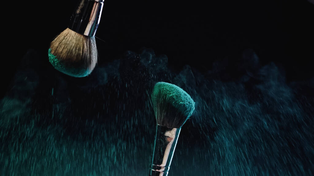 soft cosmetic brushes with turquoise holi powder splashing on black background - Photo, Image