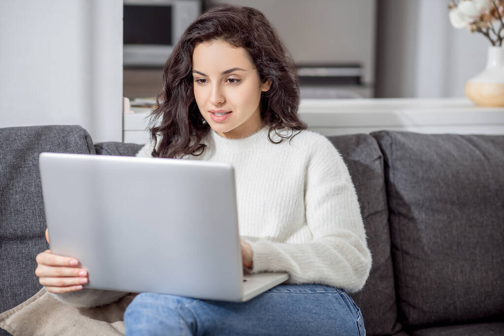 Молодая женщина в белом свитере сидит на диване с ноутбуком - Фото, изображение