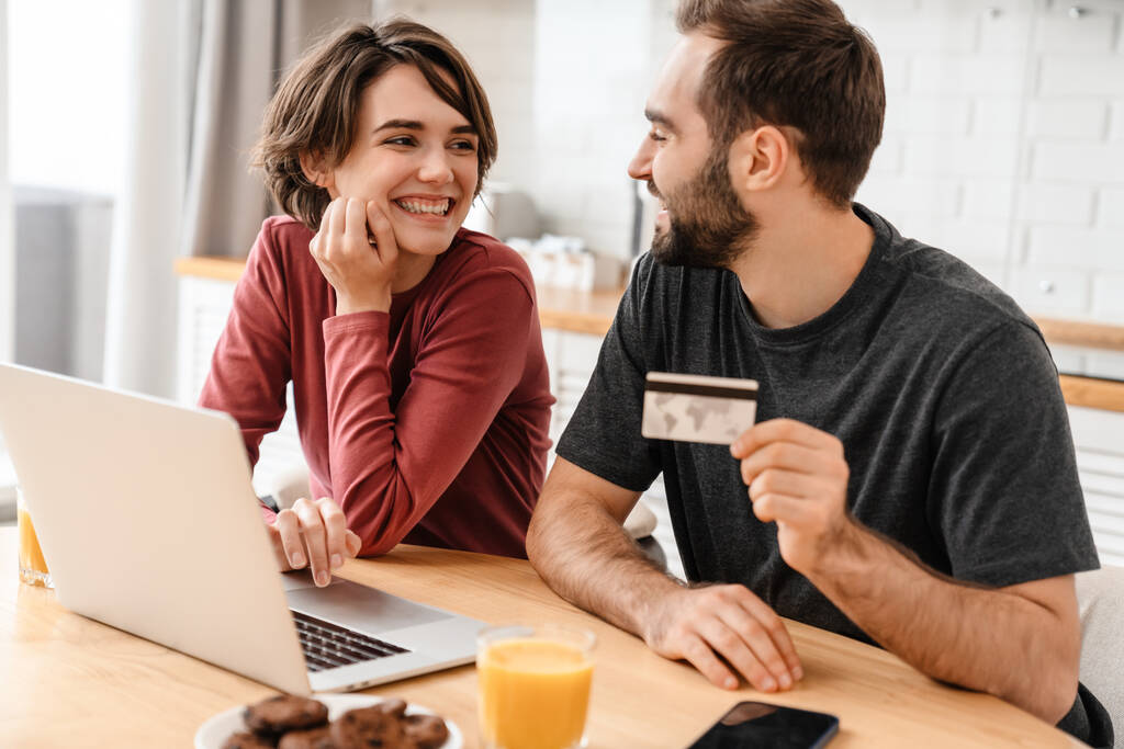 Onnellinen nuori pari käyttää kannettavaa tietokonetta ja luottokorttia kotona istuessaan - Valokuva, kuva