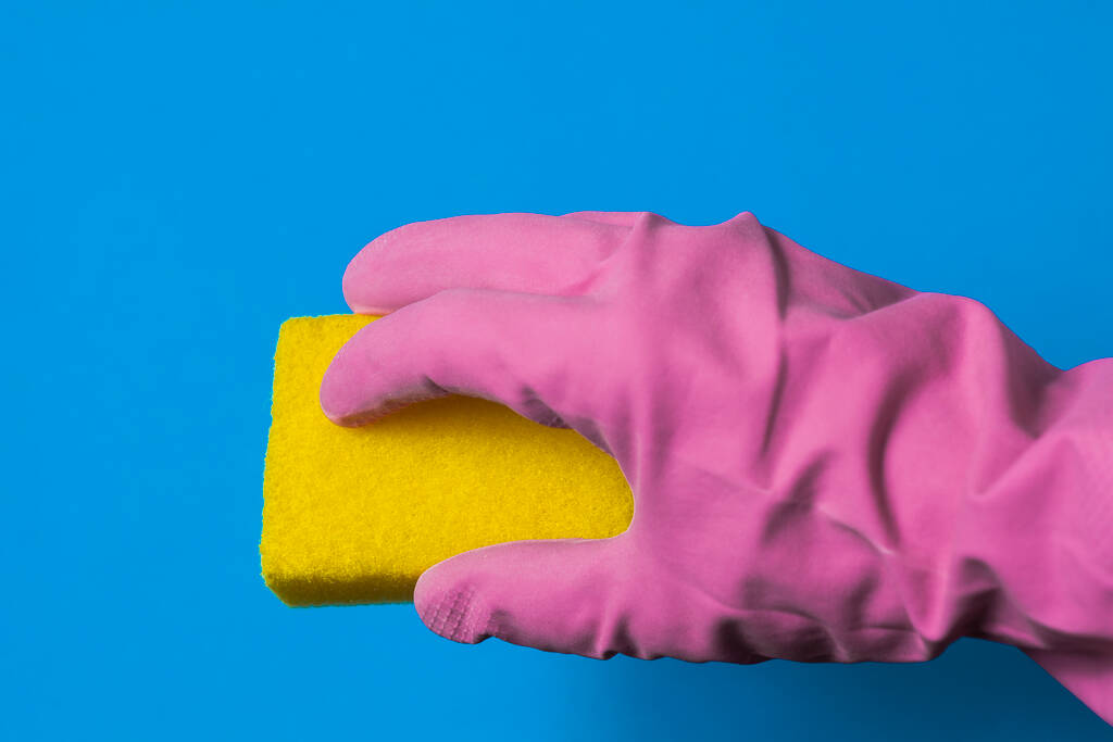 Een hand in een roze rubberen handschoen houdt een gele spons vast. Blauwe achtergrond. - Foto, afbeelding