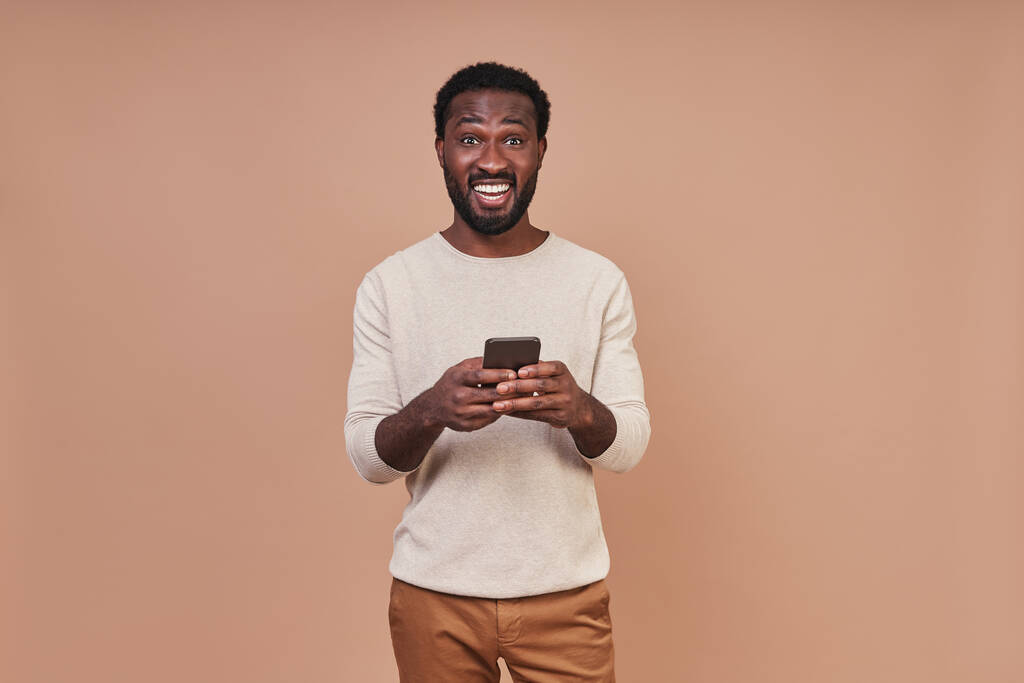 Hezký mladý Afričan v ležérní oblečení pomocí chytrého telefonu a s úsměvem - Fotografie, Obrázek