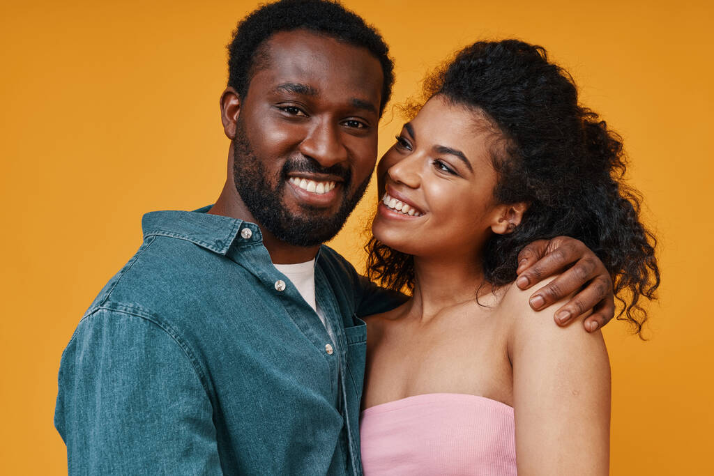 Όμορφο νεαρό ζευγάρι από την Αφρική αγκαλιάζει και χαμογελά ενώ στέκεται στο κίτρινο φόντο - Φωτογραφία, εικόνα