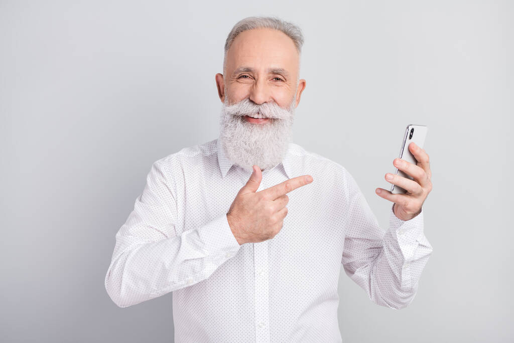 Foto van vrolijke positieve oude zakenman wijzen vinger smartphone glimlach geïsoleerd op grijze kleur achtergrond - Foto, afbeelding