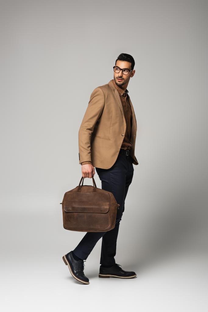 Gestor árabe bien parecido sosteniendo el bolso mientras camina sobre fondo gris - Foto, imagen