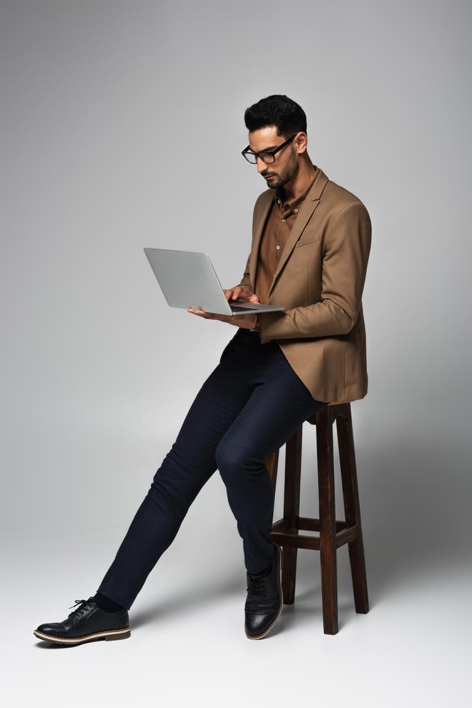 Hombre de negocios árabe usando portátil en silla sobre fondo gris - Foto, imagen