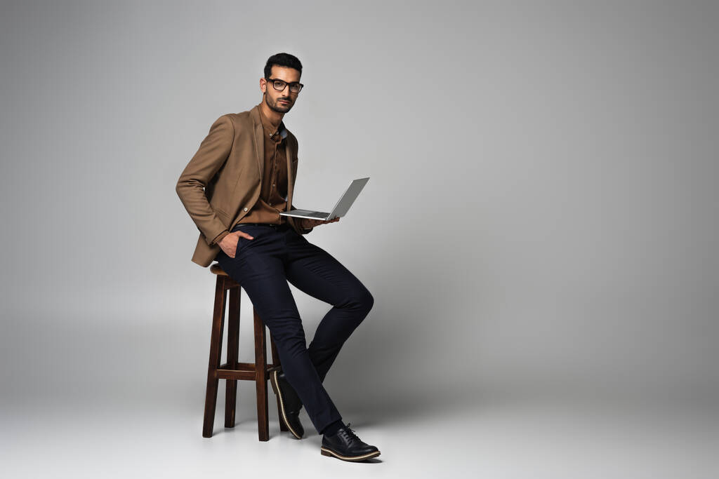 Μουσουλμάνος επιχειρηματίας με χέρι στην τσέπη και φορητό υπολογιστή κοντά καρέκλα σε γκρι φόντο - Φωτογραφία, εικόνα