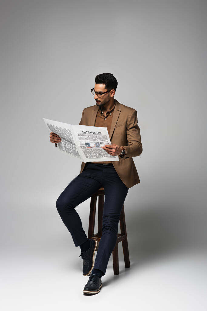 Μουσουλμάνος επιχειρηματίας που διαβάζει επιχειρηματική εφημερίδα σε καρέκλα σε γκρι φόντο - Φωτογραφία, εικόνα