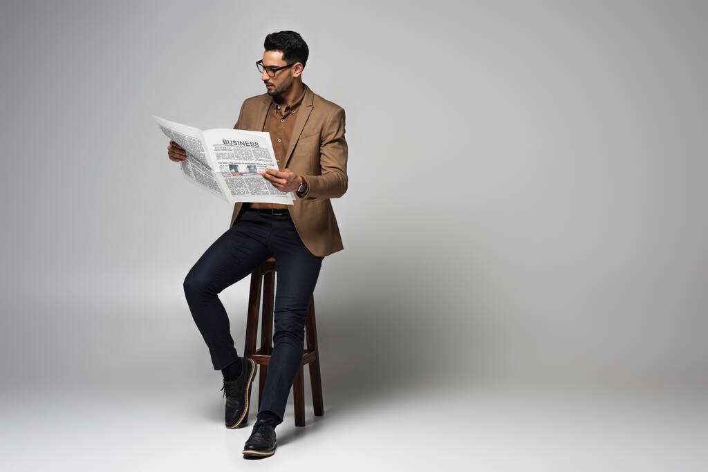Красивый мусульманский бизнесмен читает новости на стуле на сером фоне - Фото, изображение