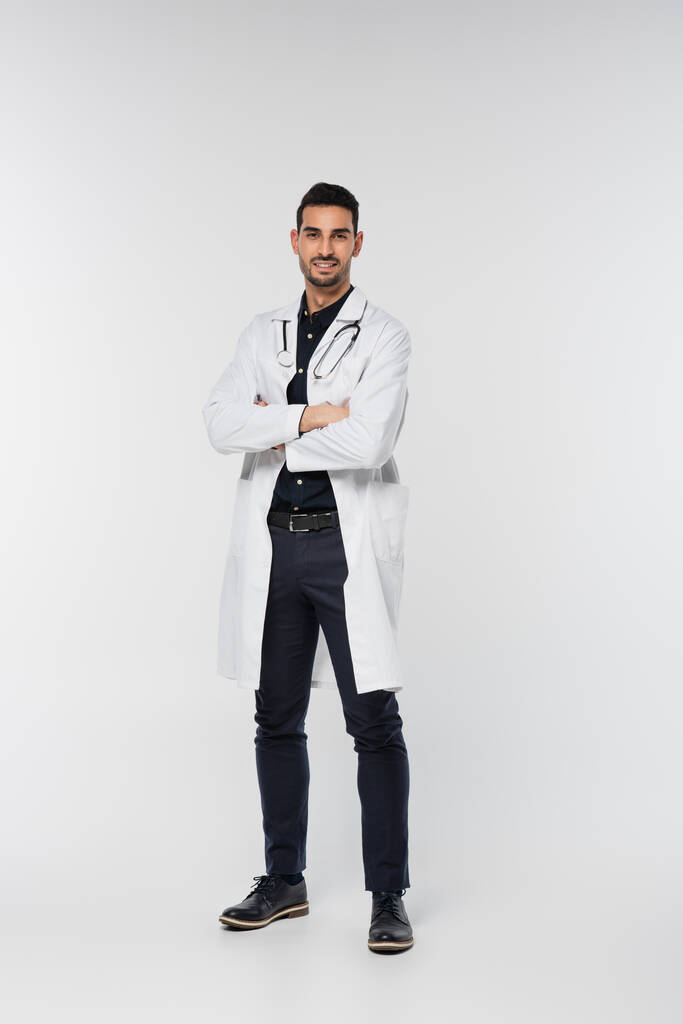 Médico árabe sonriente de pie con brazos cruzados sobre fondo gris - Foto, imagen