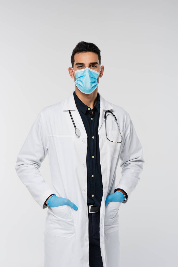 Άραβας γιατρός με ιατρική μάσκα και γάντια λατέξ κοιτάζοντας κάμερα απομονωμένη σε γκρι  - Φωτογραφία, εικόνα
