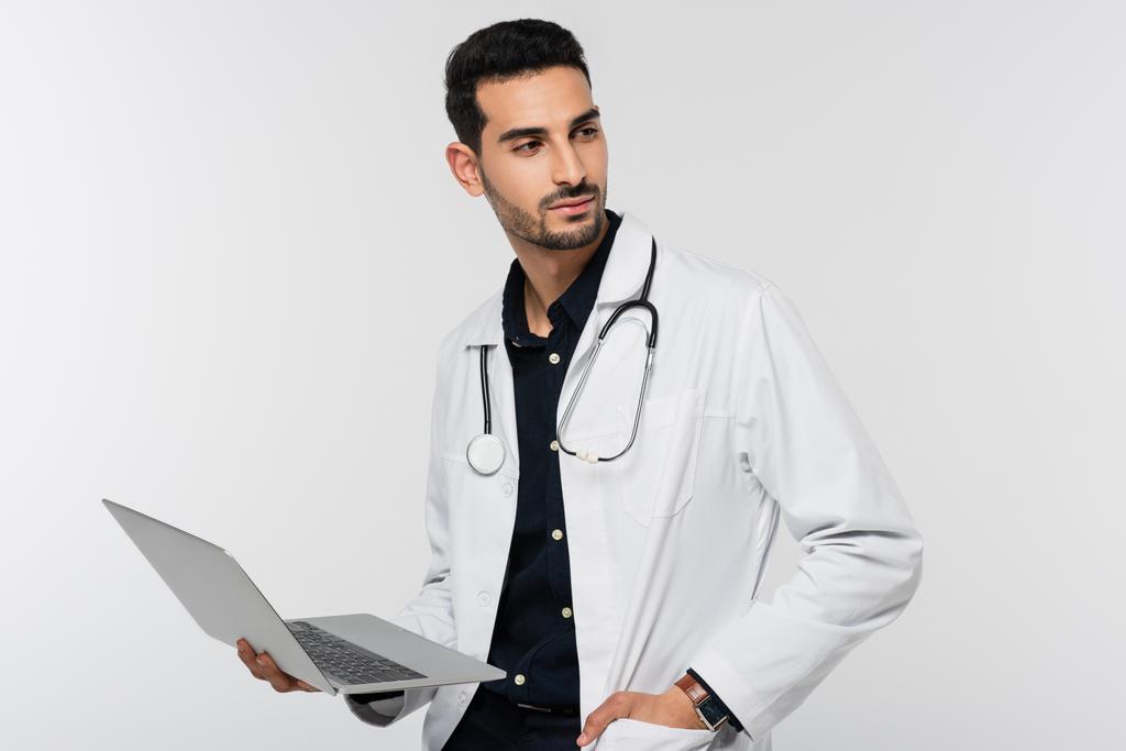 Médico árabe segurando laptop e mão no bolso de casaco branco isolado em cinza  - Foto, Imagem