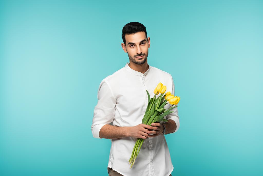 Arabský muž se žlutými tulipány hledící na kameru izolovanou na modré  - Fotografie, Obrázek