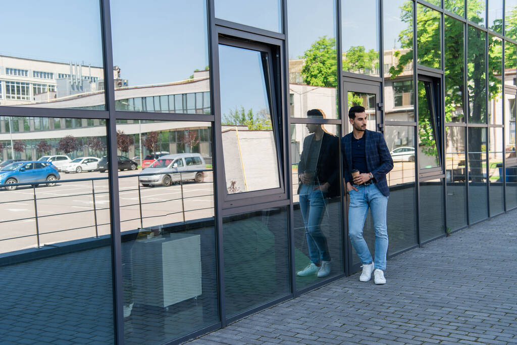 Молодой мусульманский бизнесмен держит руку в кармане и бумажный стакан рядом со зданием  - Фото, изображение