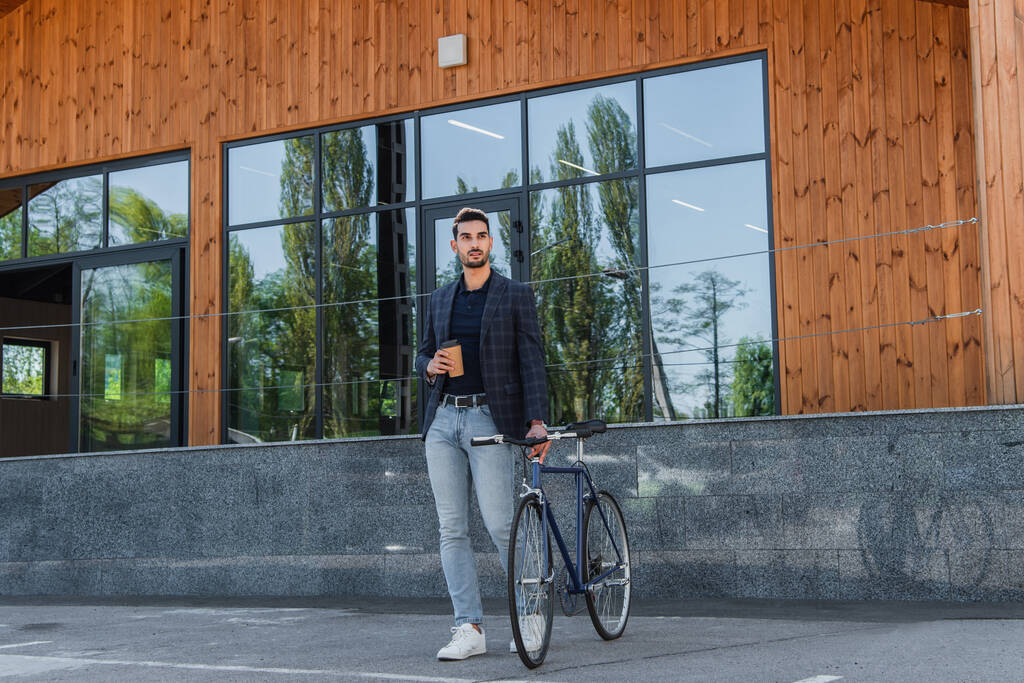 Fiatal arab menedzser kezében papír pohár közel kerékpár és épület  - Fotó, kép