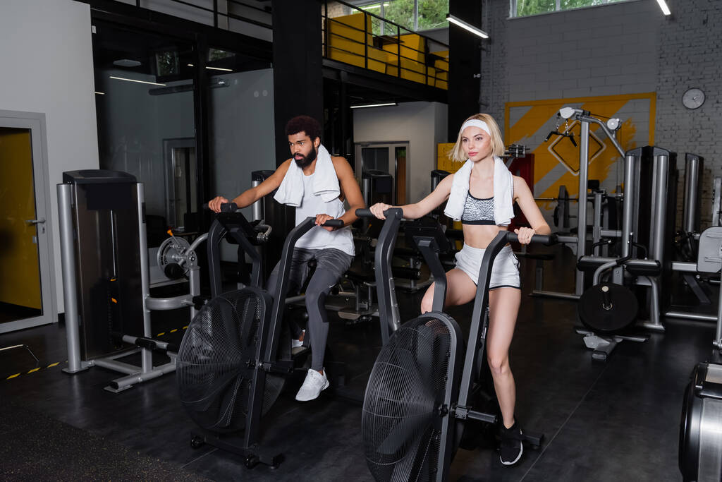 блондинка и бородатый афроамериканец тренируются на тренажерах в спортзале - Фото, изображение