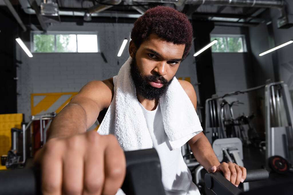 deportista afroamericano seguro de sí mismo mirando la cámara mientras hace ejercicio en el gimnasio en primer plano borroso - Foto, imagen