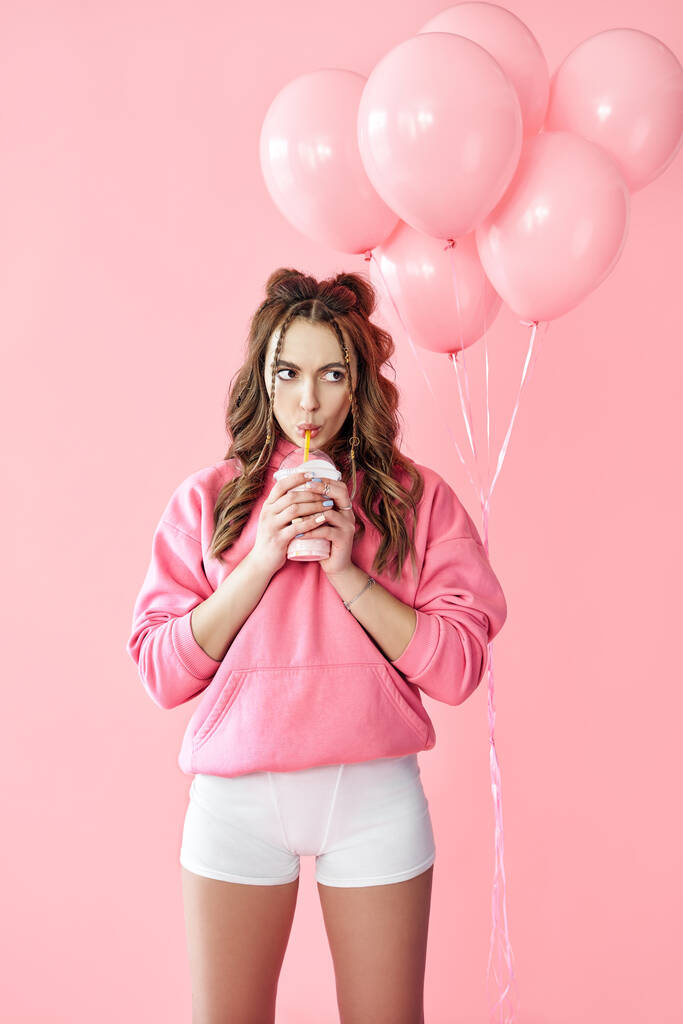 Jeune femme branchée qui boit du lait secoué sur fond de ballons roses. - Photo, image