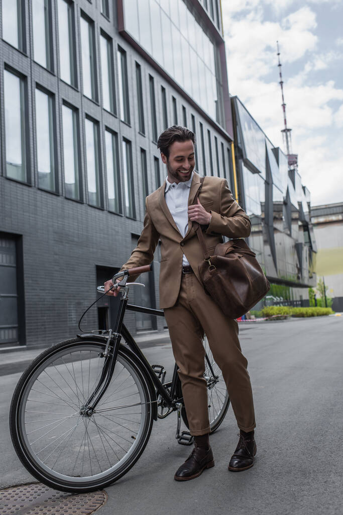 longitud completa de hombre de negocios feliz en traje con bolsa de cuero y sonriendo cerca de la bicicleta fuera  - Foto, imagen