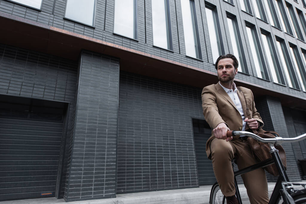 молодой бизнесмен в костюме с кожаной сумкой катается на велосипеде снаружи  - Фото, изображение