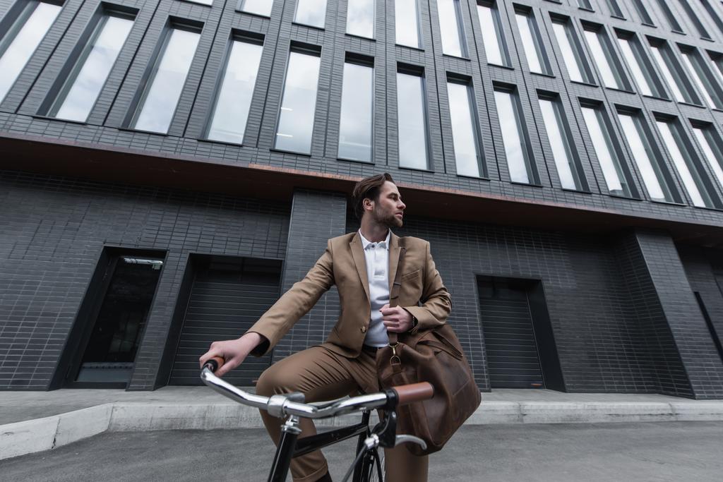 νεαρός επιχειρηματίας σε επίσημο ένδυμα με δερμάτινη καφέ τσάντα ιππασίας ποδήλατο έξω  - Φωτογραφία, εικόνα