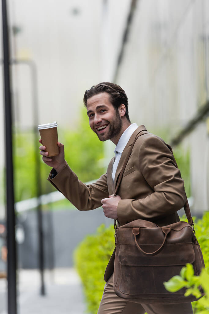 uomo d'affari sorridente in formale cinghia di tenuta di usura sulla borsa e tazza di carta vicino alla pianta offuscata  - Foto, immagini