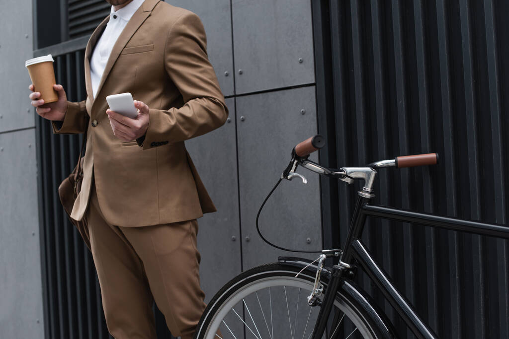 καλλιεργημένη άποψη του επιχειρηματία κρατώντας χάρτινο κύπελλο και smartphone κοντά στο ποδήλατο  - Φωτογραφία, εικόνα
