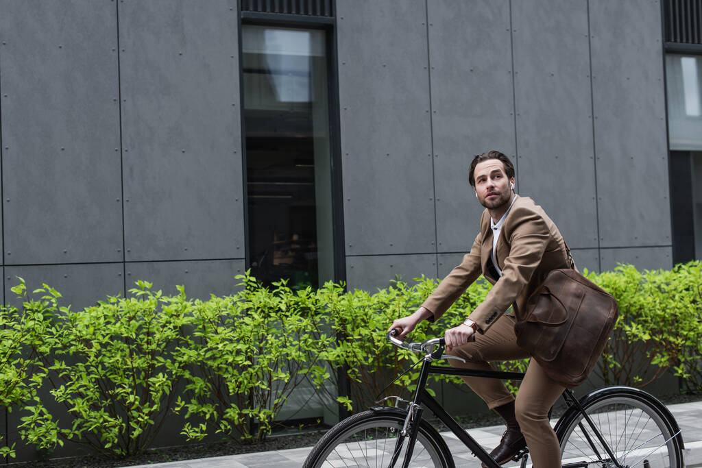 mladý podnikatel ve sluchátkách jízda na kole v blízkosti budovy  - Fotografie, Obrázek