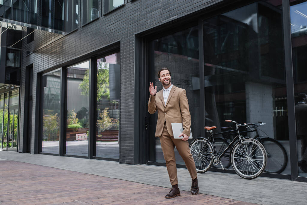 полная длина счастливый бизнесмен в наушниках размахивая рукой и держа ноутбук возле здания и велосипеда  - Фото, изображение