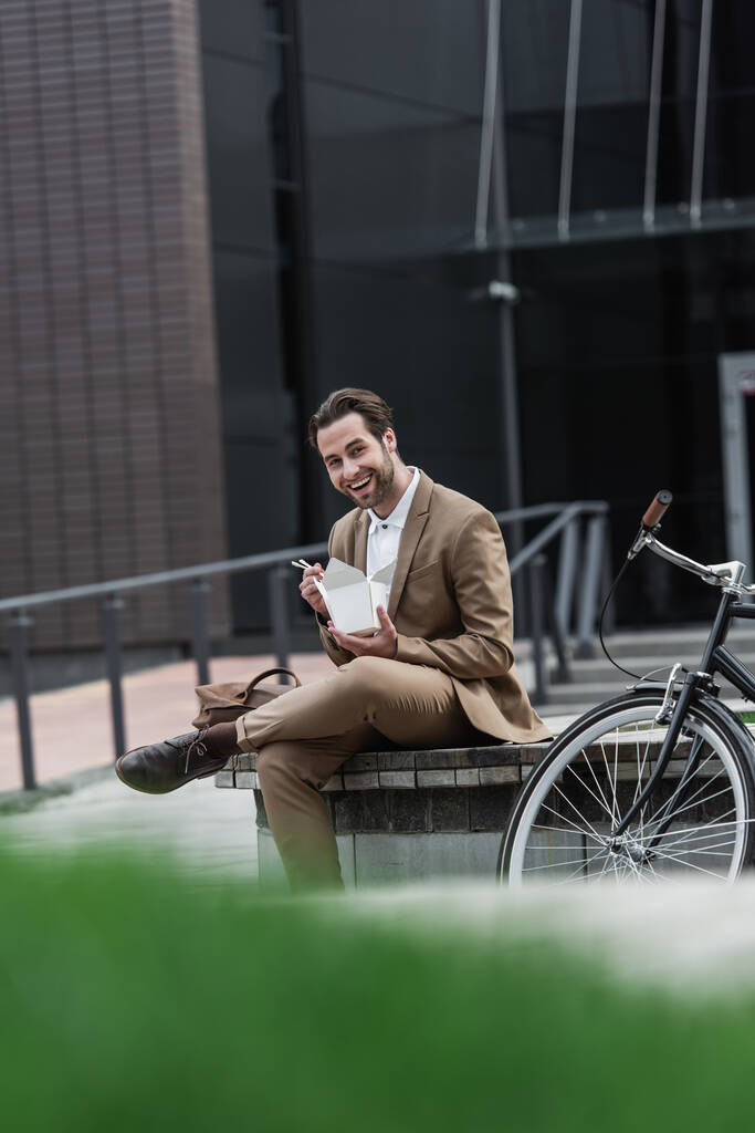 glücklicher Geschäftsmann im Anzug mit Pappschachtel mit asiatischem Essen und Essstäbchen in Fahrradnähe  - Foto, Bild